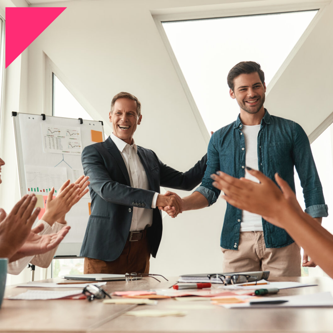 Manager shaking employees hand showing his support for a job well done in the office in front of white board with employees sitting around round desk clapping.