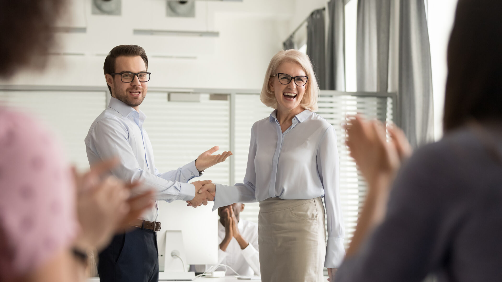 Happy manager boss praising female employee for good work and promotion. Shaking her hand and team members clapping