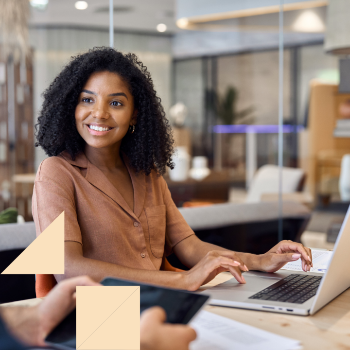 Professional woman smiling while working on a laptop during a meeting about retained search recruiting in a modern office setting, with another person holding a tablet in the foreground.