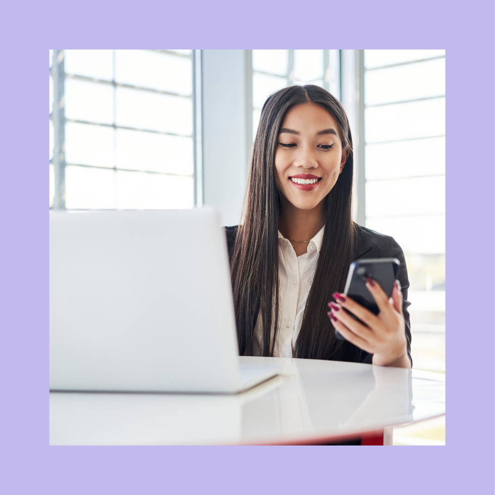 Smiling hiring manager in a business suit sitting at a desk with a laptop, checking her phone while reviewing digital recruiting trends in a modern office setting.
