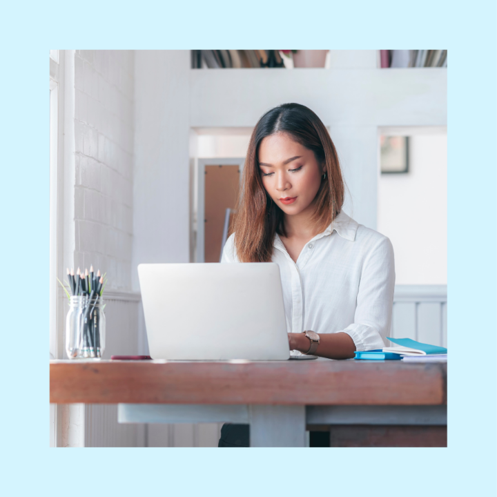 A professional woman in a white blouse working on her laptop at a wooden desk in a bright, modern home office. She appears focused, with documents and stationery neatly arranged beside her.