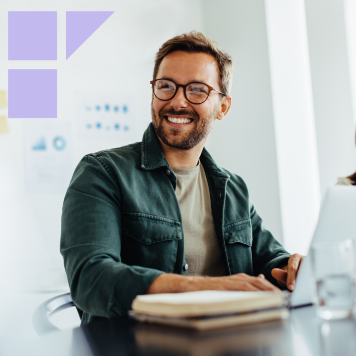 A smiling man wearing glasses and a green jacket sits at a desk in a modern office. He is using a laptop with an open notebook in front of him, appearing engaged in a positive work environment.