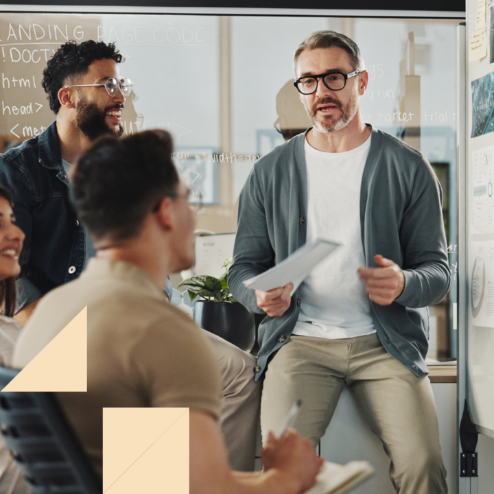 A group of diverse professionals engaged in a discussion during a workplace training session. A man in glasses and a cardigan holds a notepad while speaking, with colleagues listening attentively and taking notes in a modern office setting.