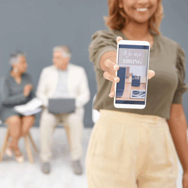 Woman holding smartphone with picture that says "we are hiring," and people waiting in the background with resumes for recruiting event