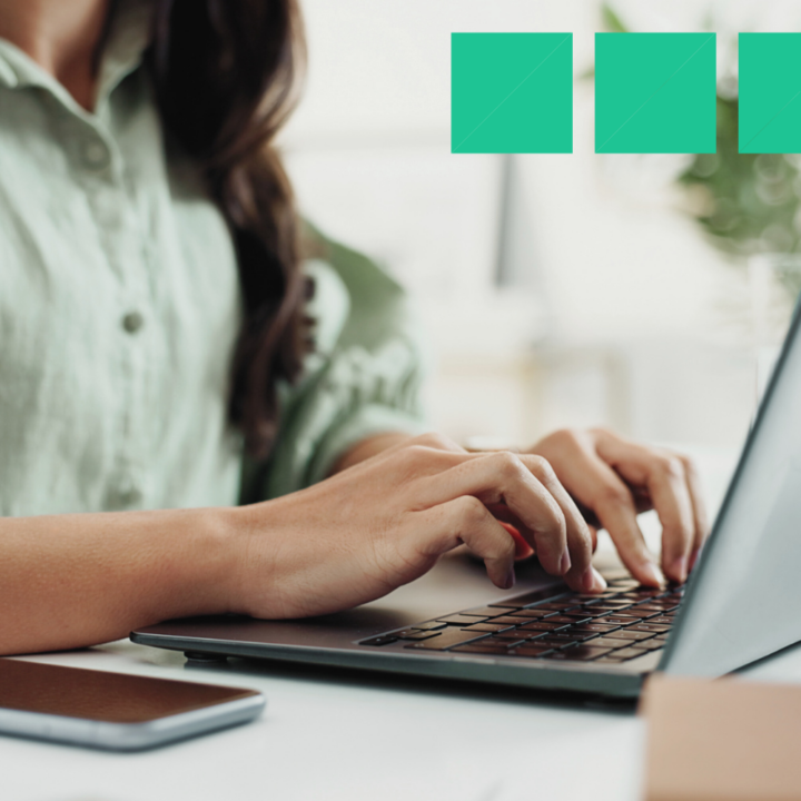 A woman in a green blouse typing on a laptop at a bright, modern workspace. A smartphone rests on the desk beside her as she focuses on writing an email.
