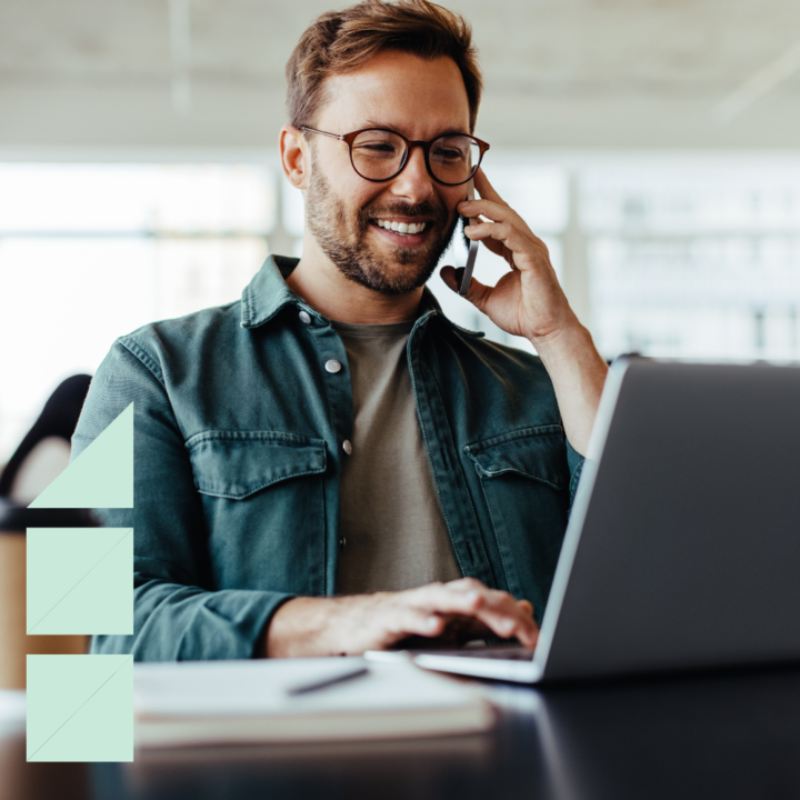 A smiling man wearing glasses and a green jacket sits at a desk in a modern office space, talking on the phone while working on his laptop. He appears to be discussing a job offer with HR.