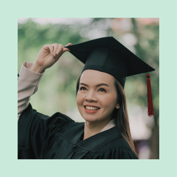 A smiling college graduate wearing a black cap and gown adjusts her graduation cap outdoors. She looks joyful and proud, celebrating her achievement in a natural setting.