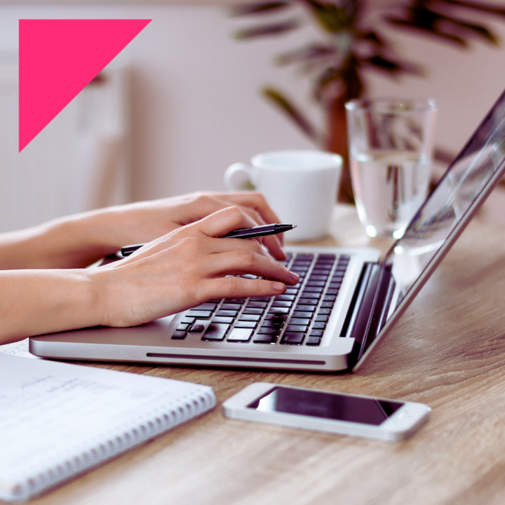 Close-up of a person's hands typing on a laptop keyboard while holding a pen. A notebook, smartphone, coffee cup, and glass of water are placed on a wooden desk.