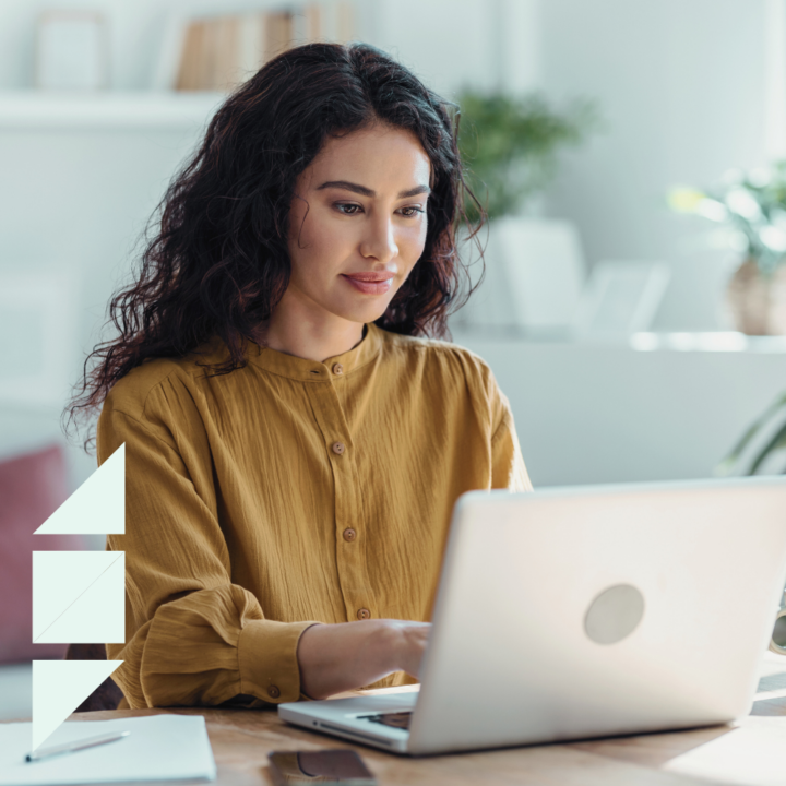 A young woman with wavy dark hair works on her laptop in a bright home office. She wears a mustard blouse and looks focused. A cover letter, pen, and phone are on the desk, with shelves and plants in the background.