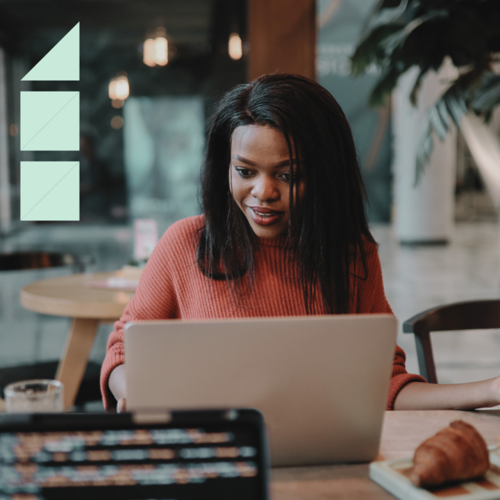A young woman in a red sweater sits at a café table, focused on her laptop with a slight smile. A croissant and a coffee cup are nearby, suggesting a relaxed yet productive atmosphere. She appears to be working on a creating a simple cover letter.