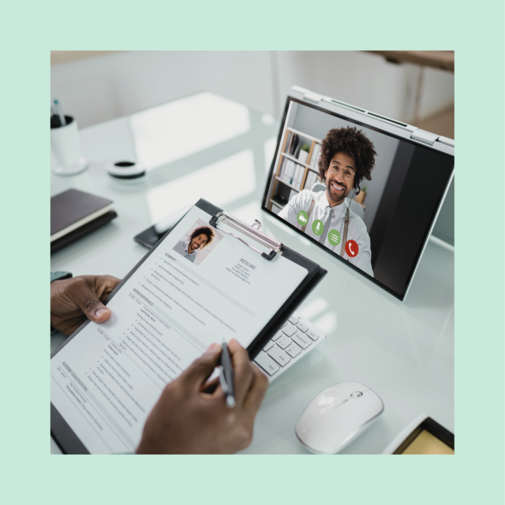 A recruiter holding a clipboard with a printed resume while conducting a virtual interview on a laptop. The candidate, a smiling man with curly hair, appears on the screen in a video call interface.