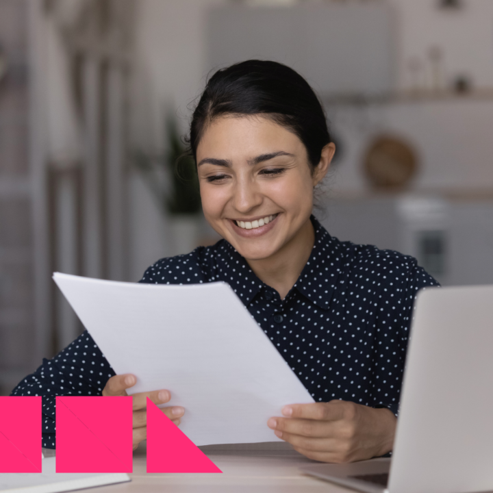 A smiling woman in a navy blue polka dot blouse sits at a desk, holding and reviewing a document, possibly a job offer. An open laptop is in front of her, and the background features a modern home setting with a kitchen area.