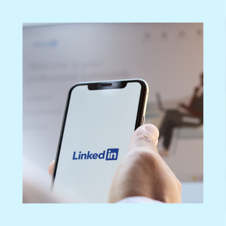 Professional holding cellphone with LinkedIn app on the screen and laptop open with the LinkedIn website open in the background