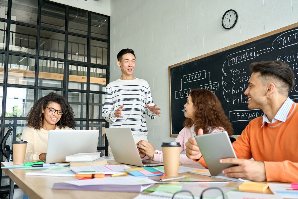 Two male and two female gen z professionals sitting around a large desk in an office with their laptops open discussing their recruitment strategy.