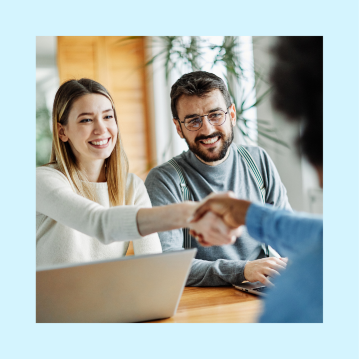 A smiling woman in a white sweater extends her hand for a handshake with a candidate, while a man in glasses and suspenders sits beside her, also smiling. A laptop is open on the table, suggesting a hiring discussion with a hiring manager.