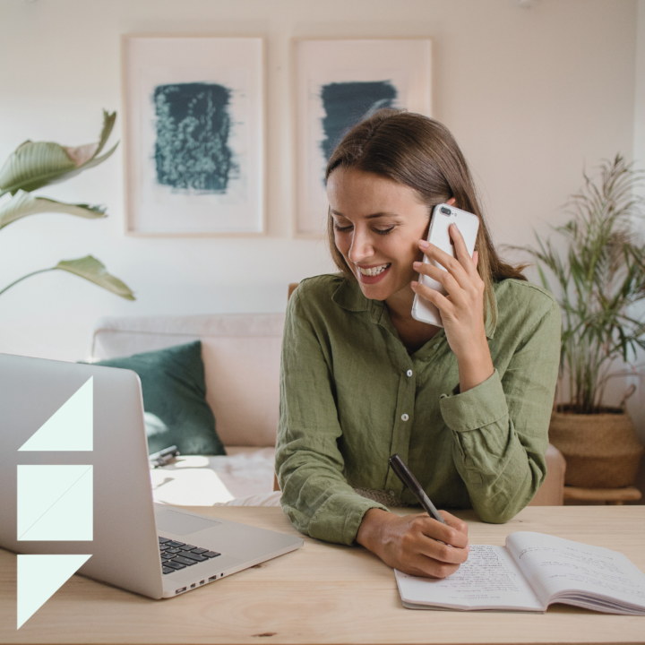 A smiling woman in a green blouse sits at a wooden desk, taking notes in a notebook while speaking on the phone. An open laptop is in front of her, and the cozy home setting includes framed artwork and plants in the background, suggesting she is engaged in a phone interview.