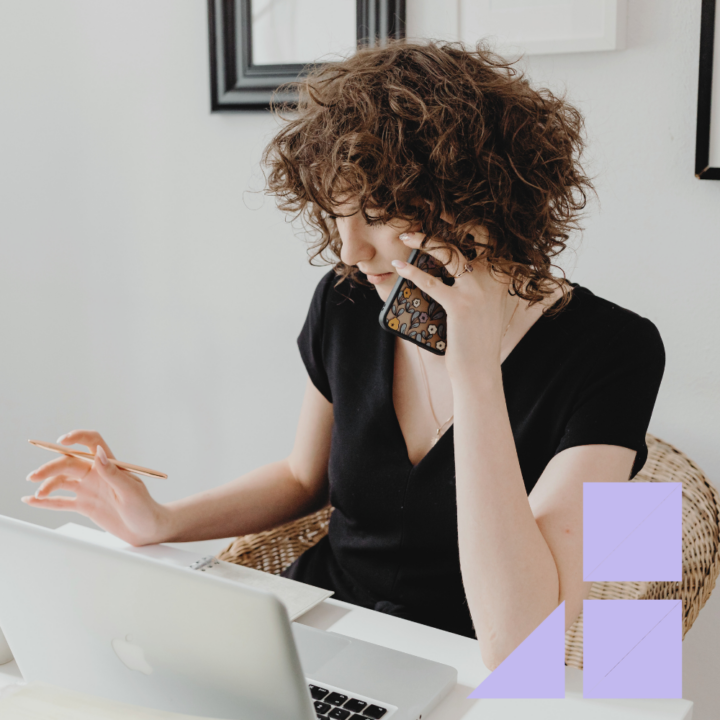 A woman with curly hair wearing a black dress is multitasking at her desk, holding a phone to her ear while taking notes with a pen. An open laptop sits in front of her as she engages in a conversation, possibly discussing contract work with a recruiter."