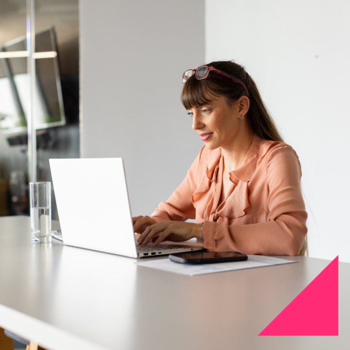A woman in a peach blouse sits at a white table, typing on her laptop with a focused expression. Her glasses rest on her head, and a smartphone and documents are nearby, suggesting she is working on a professional task, composing an apology to a customer.