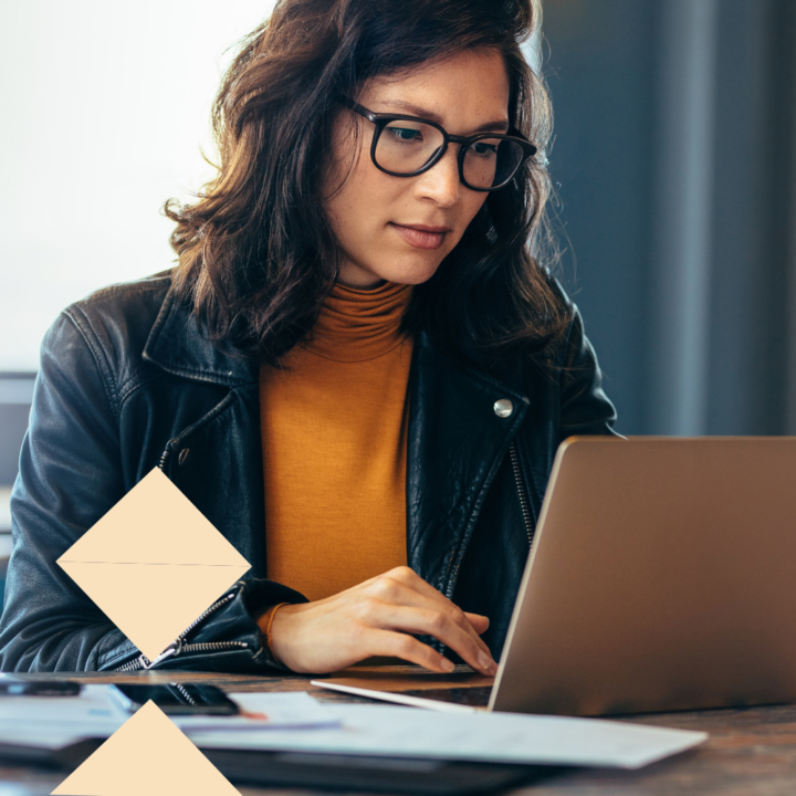 Young woman with glasses and wavy brown hair wearing a mustard-colored turtleneck and a black leather jacket, working on a laptop in a modern workspace. Papers and a smartphone are on the desk.
