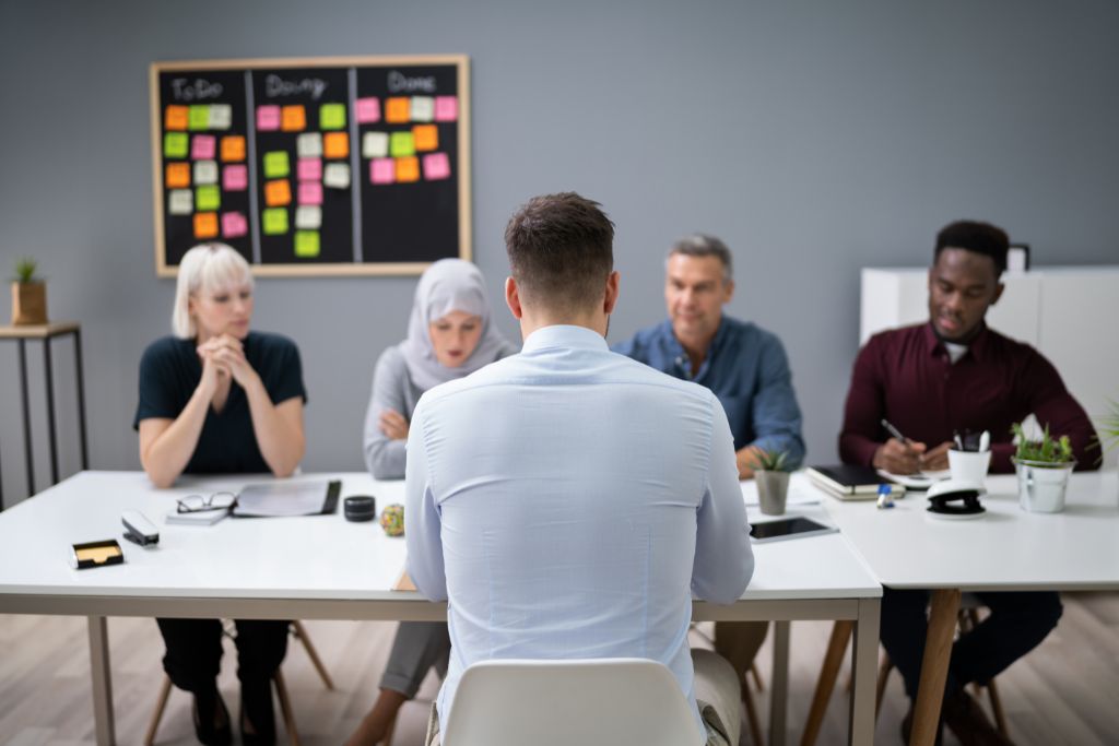 Four diverse professionals conducting a panel interview sitting at a long desk across from male job candidate. Chalkboard with colorful sticky notes behind interviewers.