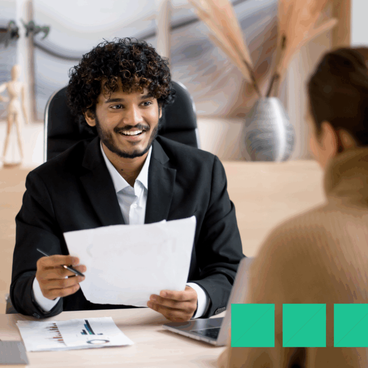 Smiling hiring manager in a black suit reviewing candidate paperwork during an in-person job interview at a modern office desk