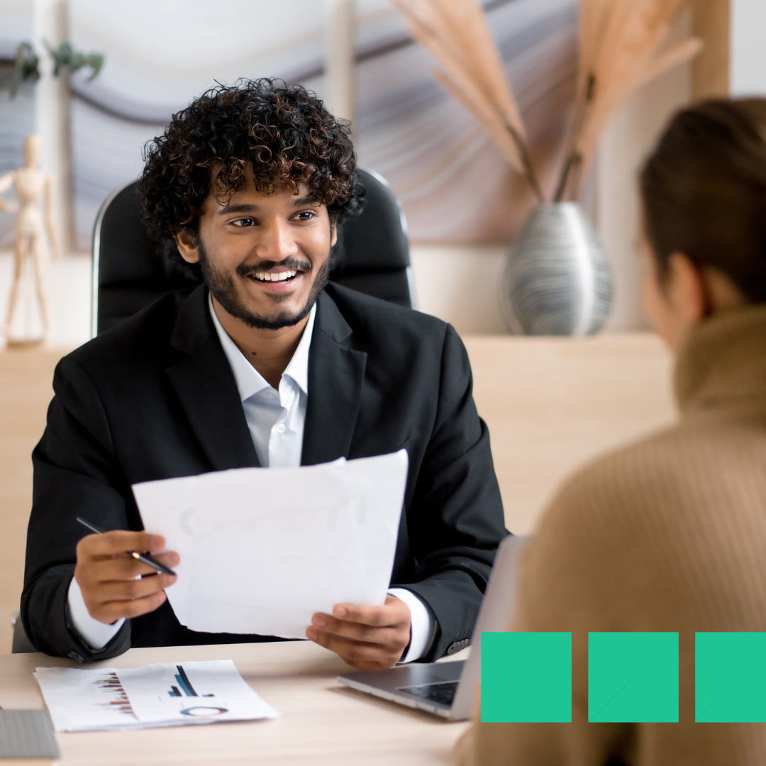 Smiling hiring manager in a black suit reviewing candidate paperwork during an in-person job interview at a modern office desk