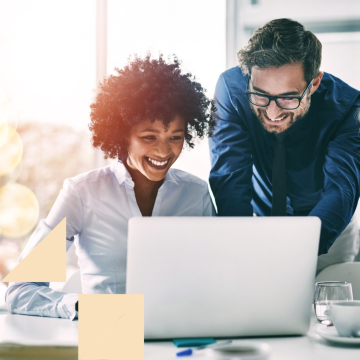 Two colleagues smiling while reviewing information on a laptop during a collaborative meeting in a bright office, suggesting teamwork, mentoring, or strategic planning in a professional workplace.