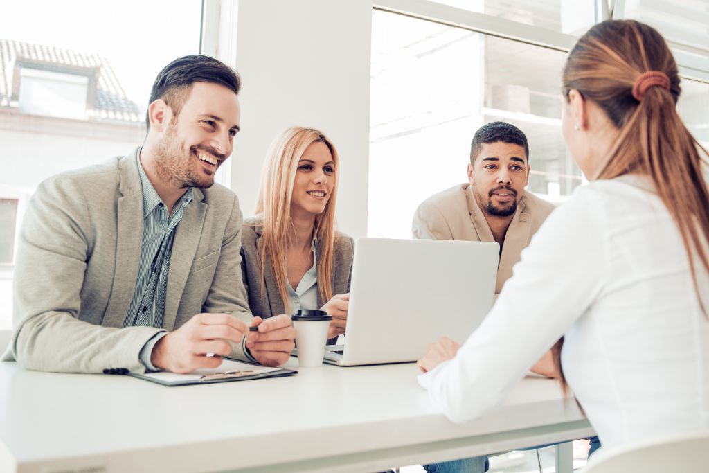 Structured job interview with one female candidate and three interviewers sitting around a desk with one laptop open and someone taking notes