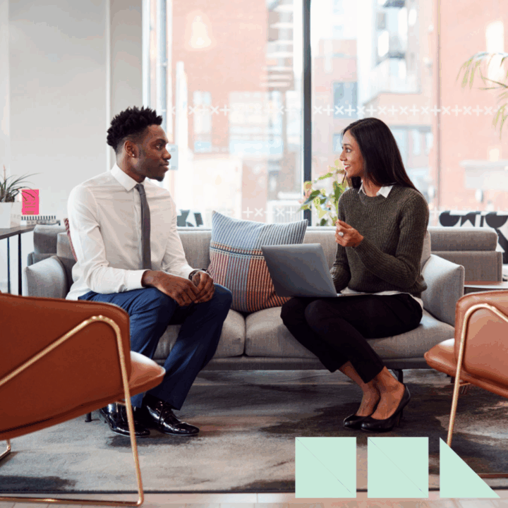A hiring manager and a job candidate sit across from each other in a modern office during an interview. The manager, holding a laptop, listens attentively while the candidate in a white shirt and tie responds. The setting includes large windows, contemporary furniture, and natural light, conveying a professional yet relaxed atmosphere.