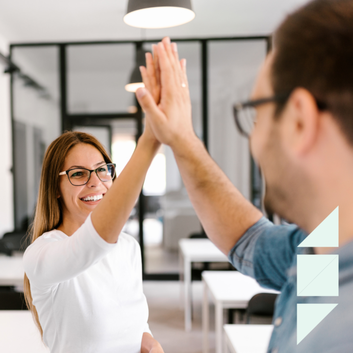Two coworkers in a modern office exchanging a high-five, smiling and celebrating a success. The background features glass walls and desks, reflecting a collaborative work environment.