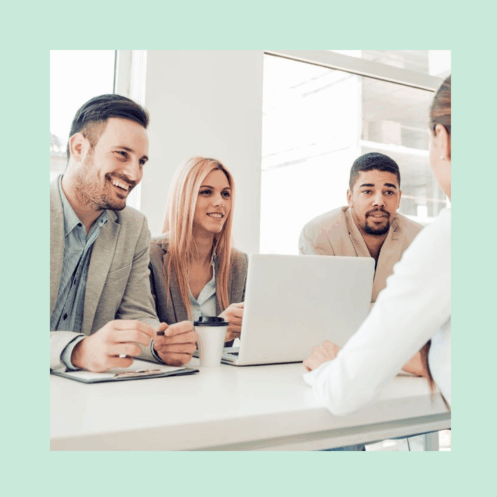 Structured job interview with one female candidate and three interviewers sitting around a desk with one laptop open and someone taking notes