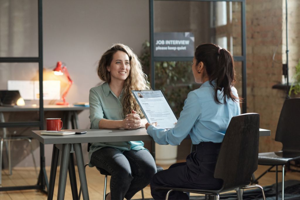 Rear view of young woman presenting her resume to director while sitting with her at table during job interview stage of candidate journey