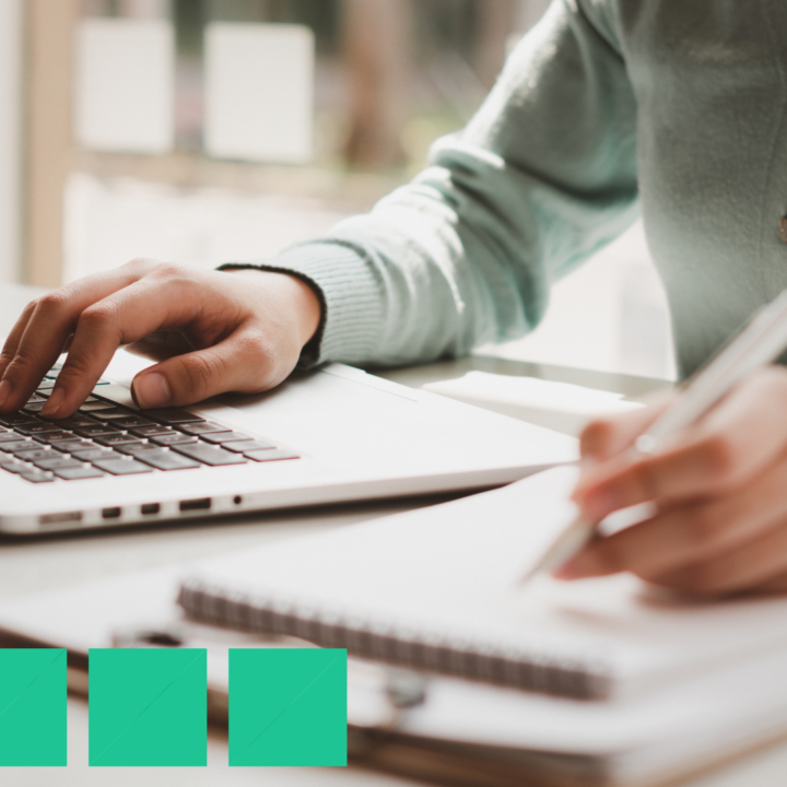 Close-up of a person working at a laptop while writing in a notebook, reviewing and checking off items on a new hire onboarding checklist at a bright, modern desk.
