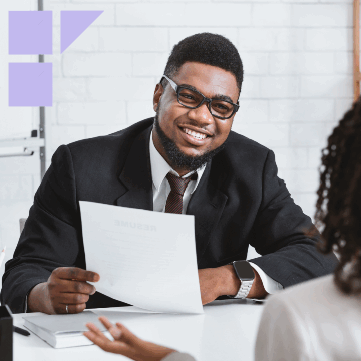 A smiling hiring manager in a suit holds a resume during an interview, seated at a desk across from a candidate. The setting is a bright, modern office with a white brick wall in the background.