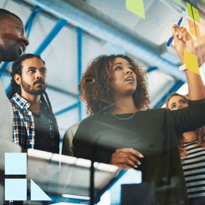A diverse group of creative professionals collaborates in a modern office. A woman writes notes on a glass board while teammates look on attentively, brainstorming ideas together.