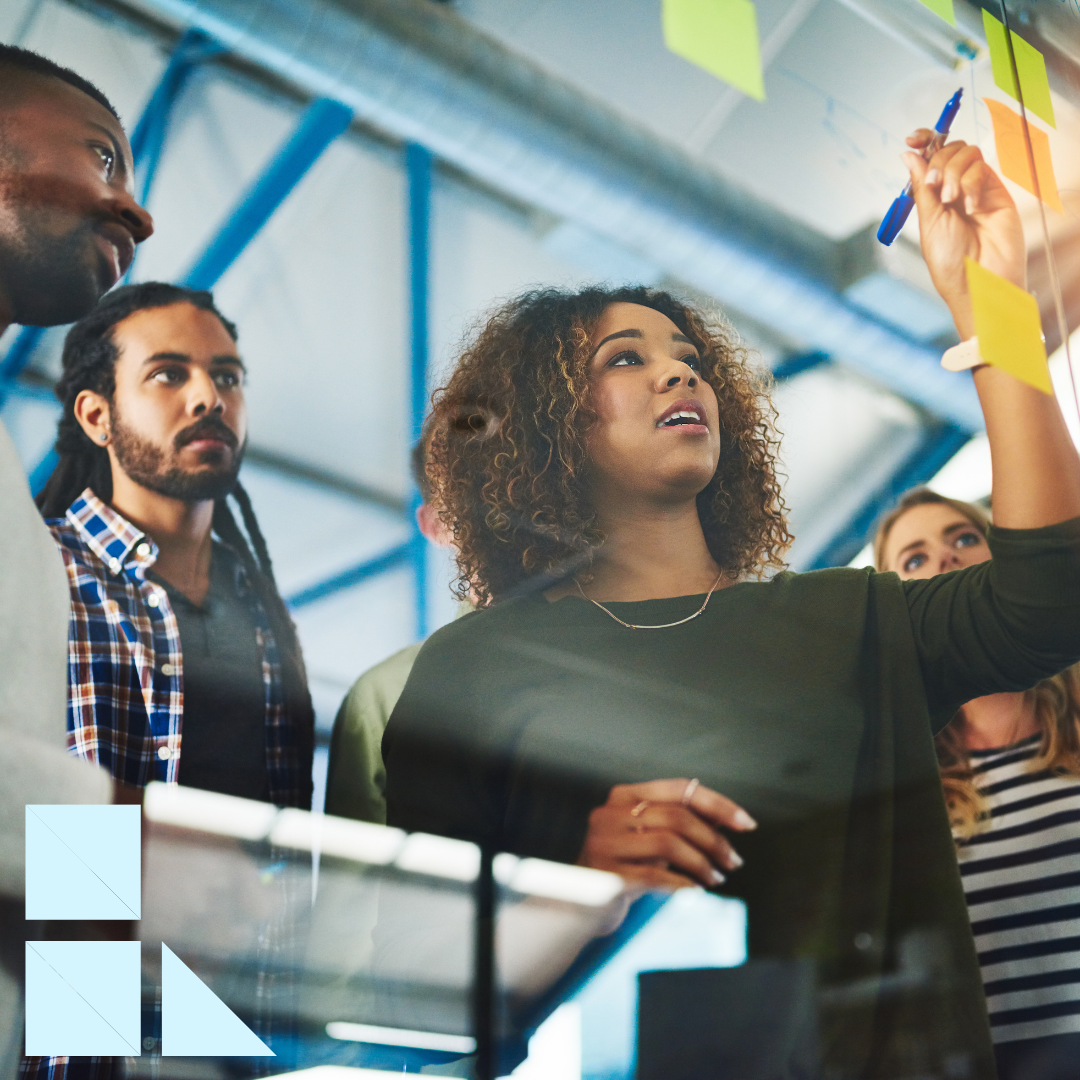 A diverse group of creative professionals collaborates in a modern office. A woman writes notes on a glass board while teammates look on attentively, brainstorming ideas together.