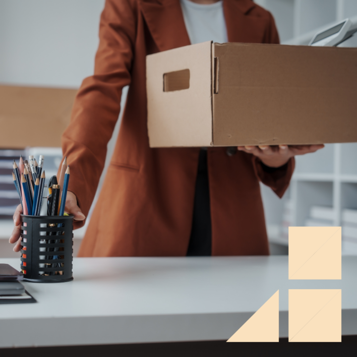 A woman wearing a brown blazer is seen packing up their belongings in an office. They are holding a cardboard box filled with items while reaching for a pen holder on the desk, symbolizing resignation or departure from the workplace.