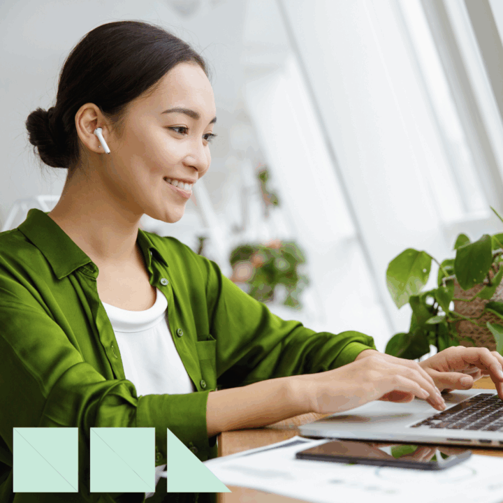 Smiling recruiter wearing a green blouse and wireless earbuds works on a laptop in a bright, modern office space with plants, representing innovative candidate sourcing techniques.