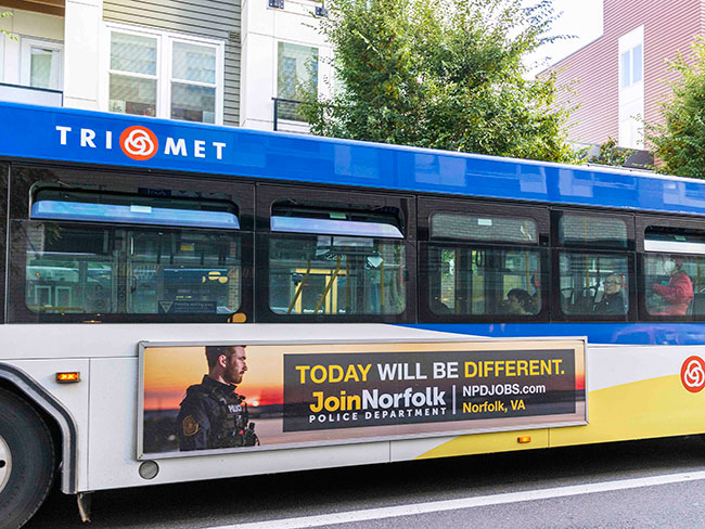 A city bus with a blue and white TriMet logo features a side advertisement for the Norfolk Police Department. The ad includes a photo of a police officer and bold yellow and white text that reads, "TODAY WILL BE DIFFERENT. Join Norfolk Police Department." It also includes the website "NPDJOBS.com" and the location "Norfolk, VA." The bus is parked on a city street in front of residential buildings.