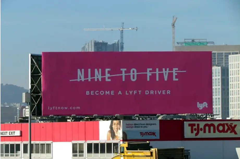 A large pink billboard with the text "NINE TO FIVE" crossed out and the words "BECOME A LYFT DRIVER" below it. The billboard promotes Lyft and includes the URL "lyftnow.com". It is situated above a T.J.Maxx store in an urban area with high-rise buildings and construction cranes in the background.