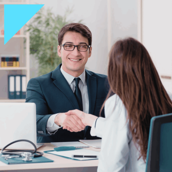 A smiling recruiter in a suit shakes hands with a female healthcare professional during a job interview in an office setting. A stethoscope and medical documents are visible on the desk, symbolizing healthcare recruiting.