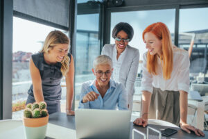 Three business woman in an office hovering over another professional during her returnship and showing her how to complete a task on her laptop