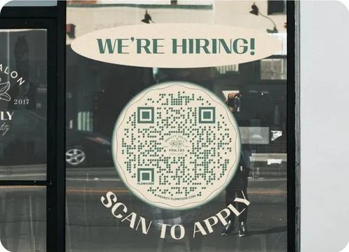 A storefront window displays a hiring sign with the text "WE'RE HIRING!" above a large circular QR code. Below the code, it reads "SCAN TO APPLY" in bold letters. The reflection of the street and a person taking the photo can be seen in the glass.