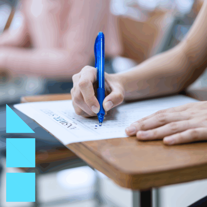 A close-up of a person’s hand holding a blue pen and filling out a multiple-choice answer sheet for a personality test on a desk. The background is blurred, showing other students sitting at desks.