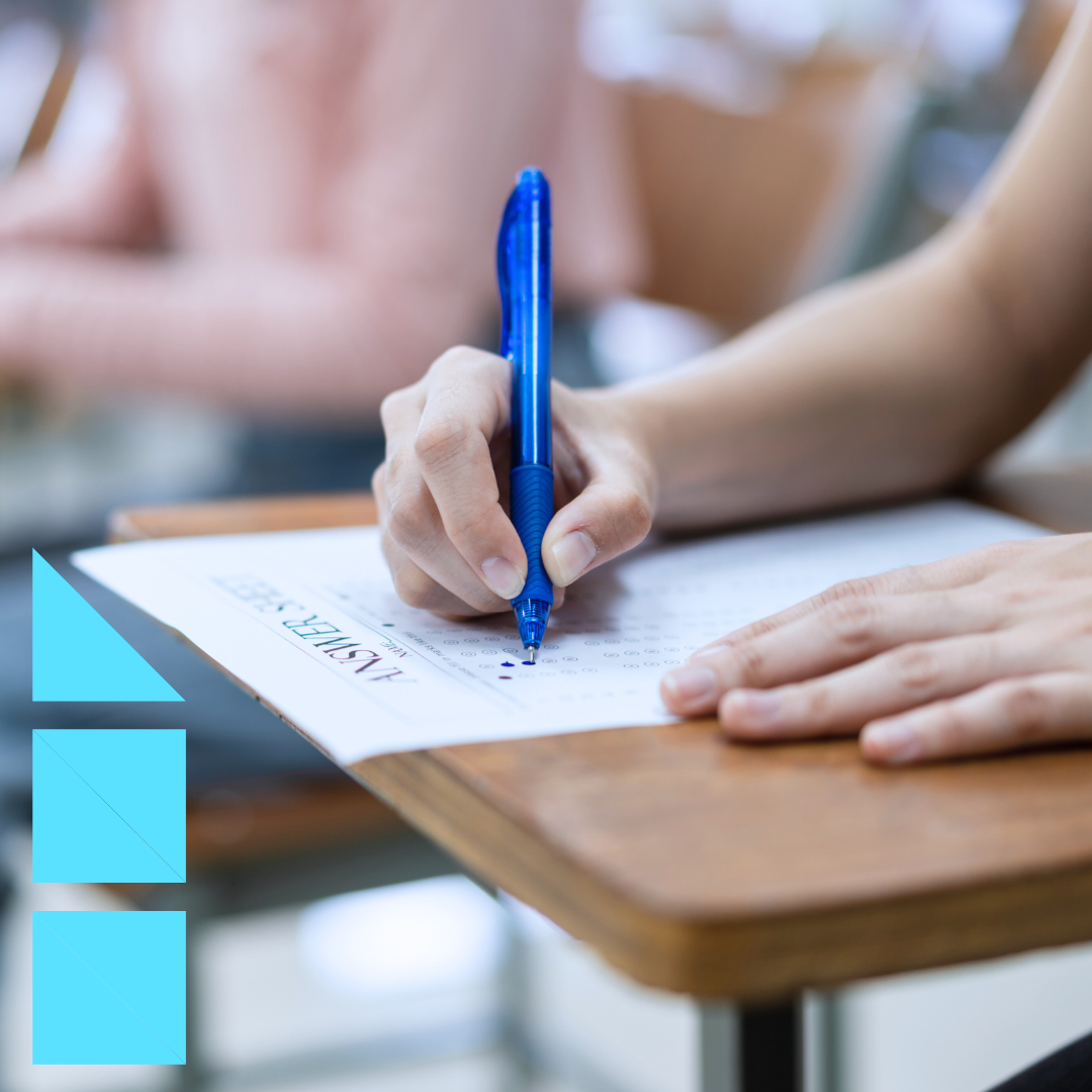 A close-up of a person’s hand holding a blue pen and filling out a multiple-choice answer sheet for a personality test on a desk. The background is blurred, showing other students sitting at desks.