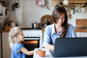 Working from home mom typing on her laptop in her kitchen sitting next to her toddler daughter
