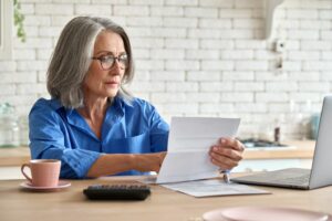 Woman professional in her seventies sitting at her kitchen table with a cup of coffee, calculator, and laptop holding and reviewing a piece of paper which is her retirement announcement.