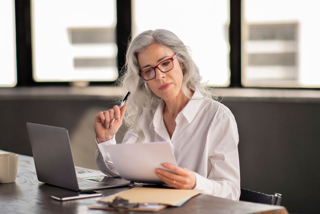Woman wearing a white blouse and red glasses sitting at her desk with a pen in her hand and holding a physical copy of her return to workforce cover letter