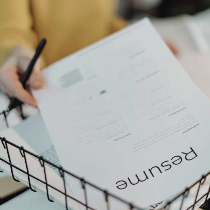 reviewing a modern, professionally designed resume placed in a wire tray. The person, wearing a yellow sweater, holds a pen while evaluating the document. The resume layout includes bold headings and structured sections.