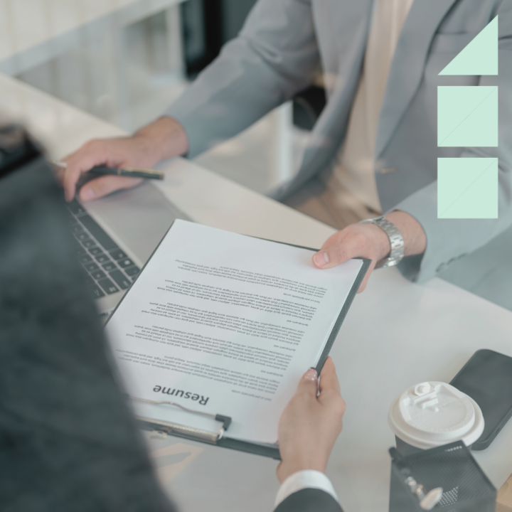 A candidate handing over a resume in a professional interview setting. The recruiter, dressed in a suit, is reviewing the document while working on a laptop. A coffee cup and office supplies are visible on the desk, creating a business-like atmosphere.